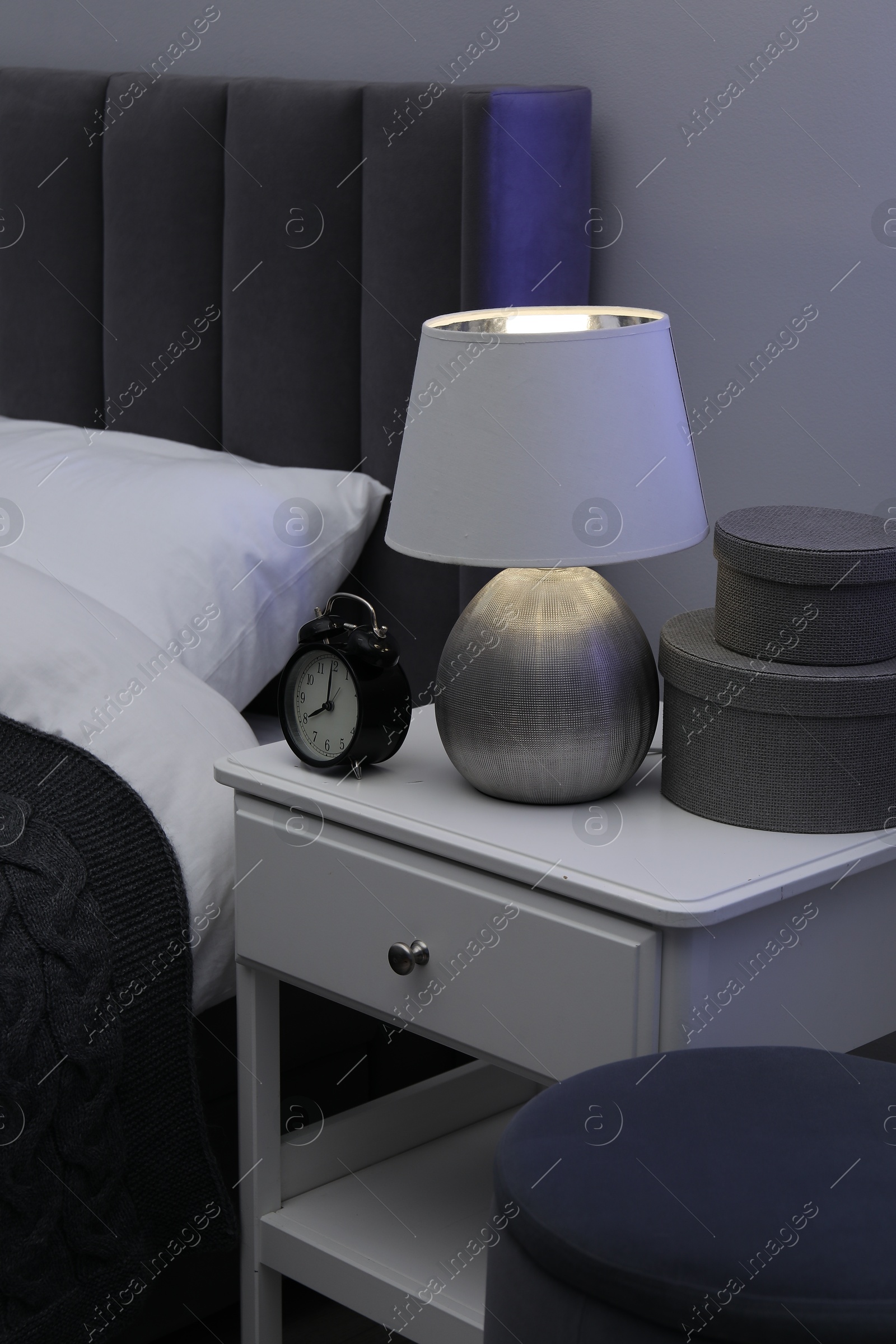 Bedside table with lamp, alarm clock and decor near bed indoors Photo of Bedside table with lamp, alarm clock and decor near bed indoors
