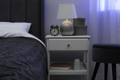 Photo of Bedside table with lamp, alarm clock and decor near bed indoors