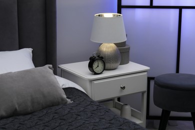 Photo of Bedside table with lamp, alarm clock and decor near bed indoors