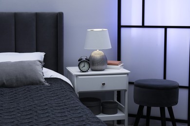 Photo of Bedside table with lamp, alarm clock and books near bed indoors