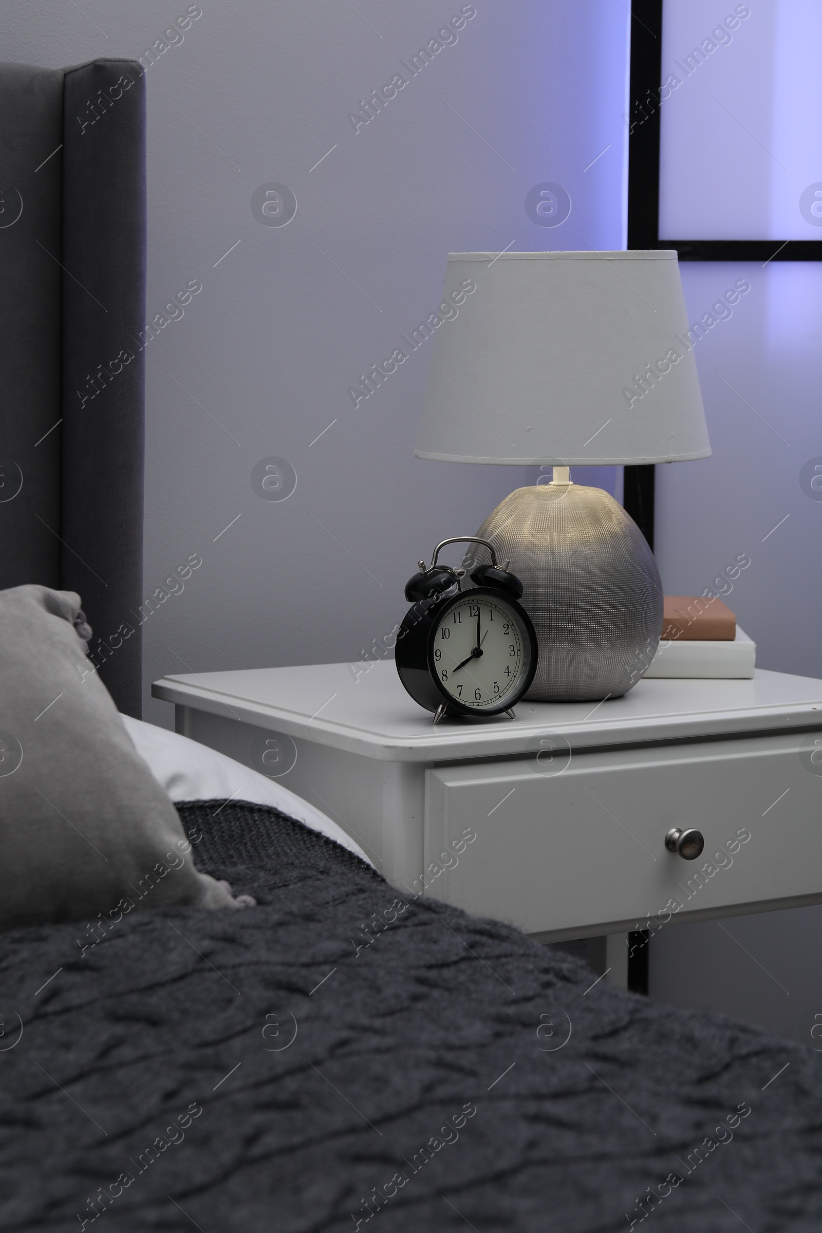Bedside table with lamp, alarm clock and books near bed indoors Photo of Bedside table with lamp, alarm clock and books near bed indoors
