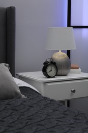 Photo of Bedside table with lamp, alarm clock and books near bed indoors