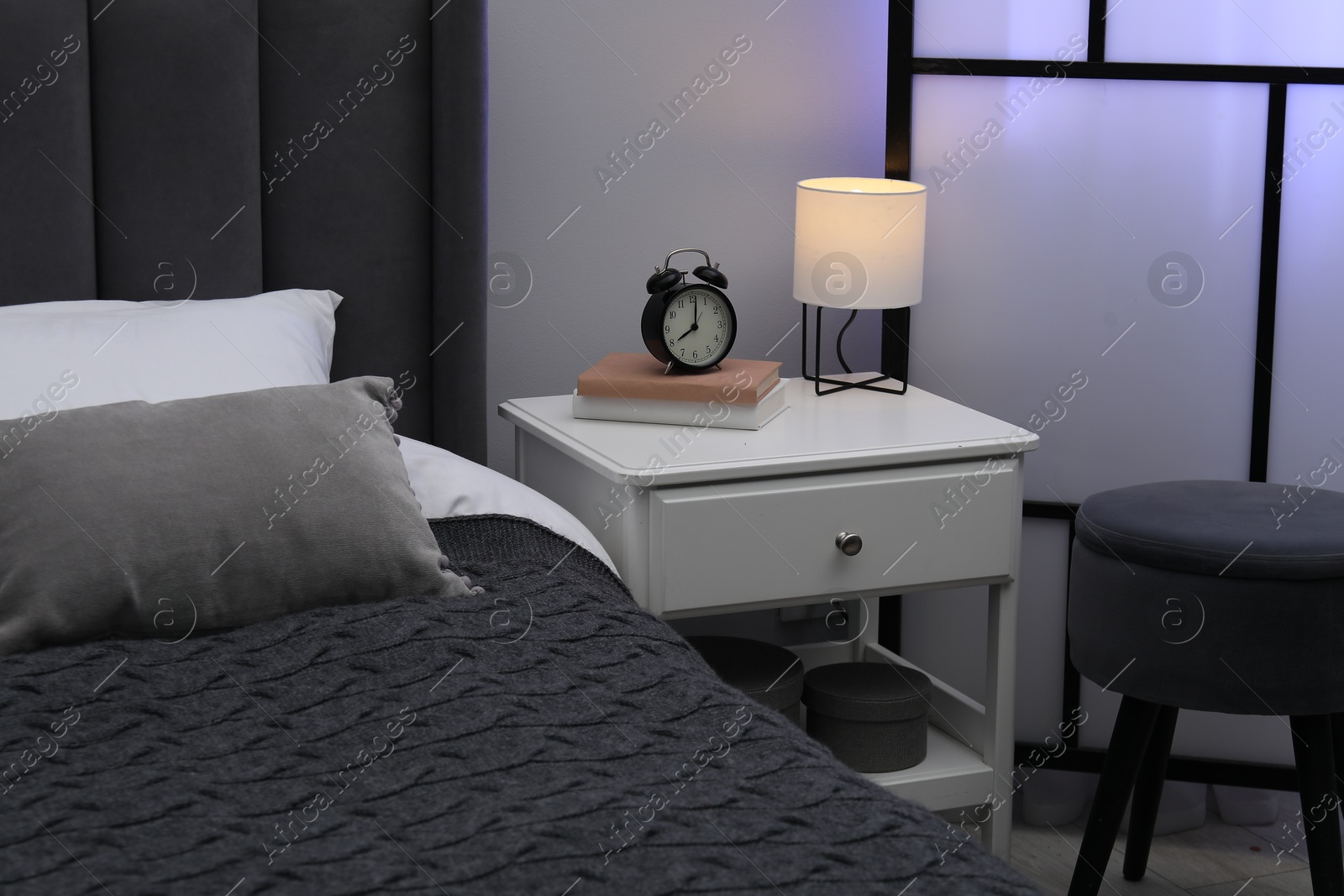 Bedside table with lamp, alarm clock and books near bed indoors Photo of Bedside table with lamp, alarm clock and books near bed indoors