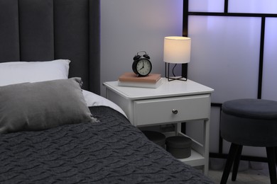 Photo of Bedside table with lamp, alarm clock and books near bed indoors