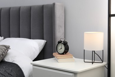 Photo of Bedside table with lamp, alarm clock and books near bed indoors