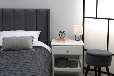 Photo of Bedside table with lamp, alarm clock and books near bed indoors