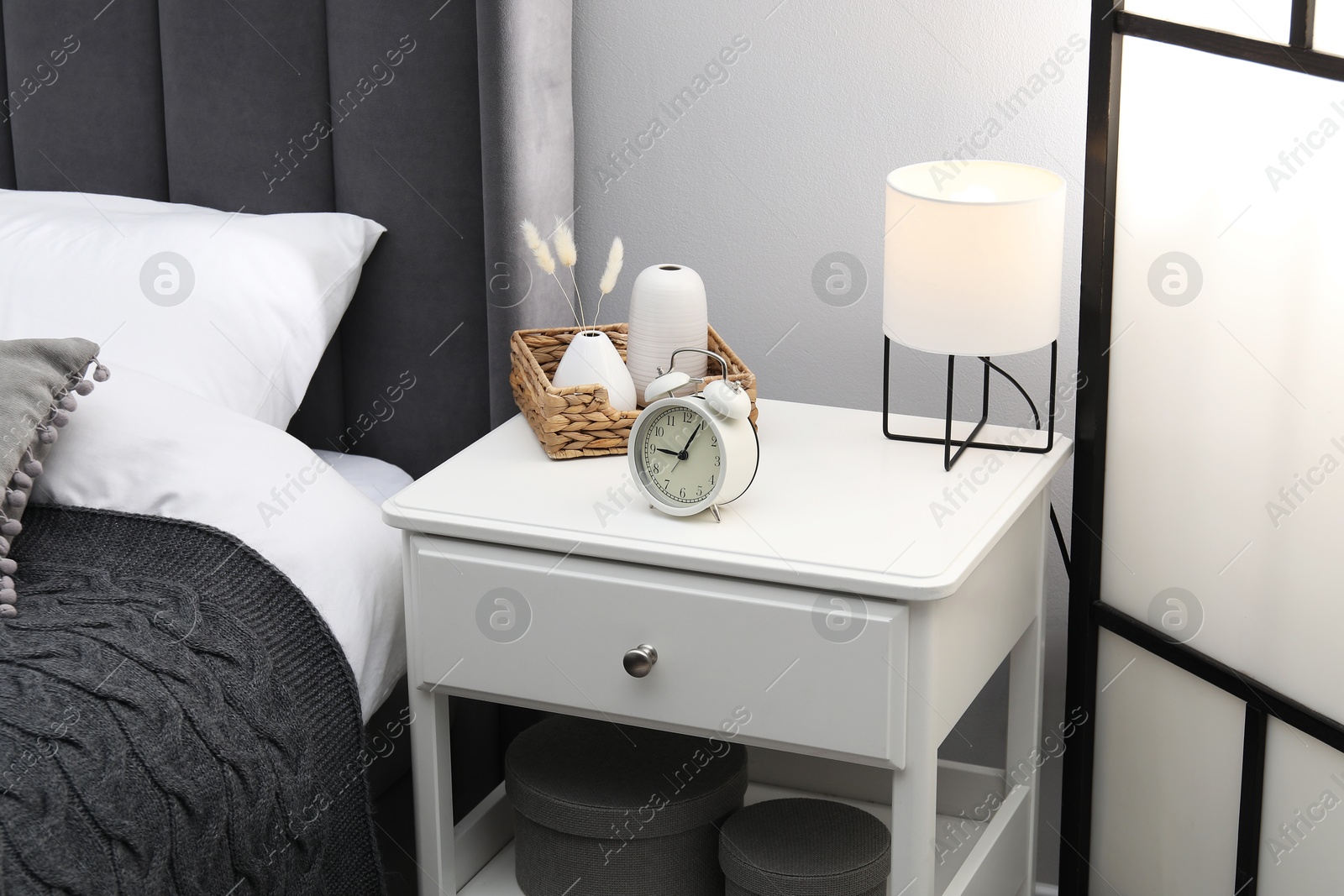 Photo of Bedside table with lamp, alarm clock and decor near bed indoors