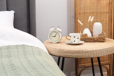 Photo of Side table with alarm clock, cup of drink and decor near bed indoors
