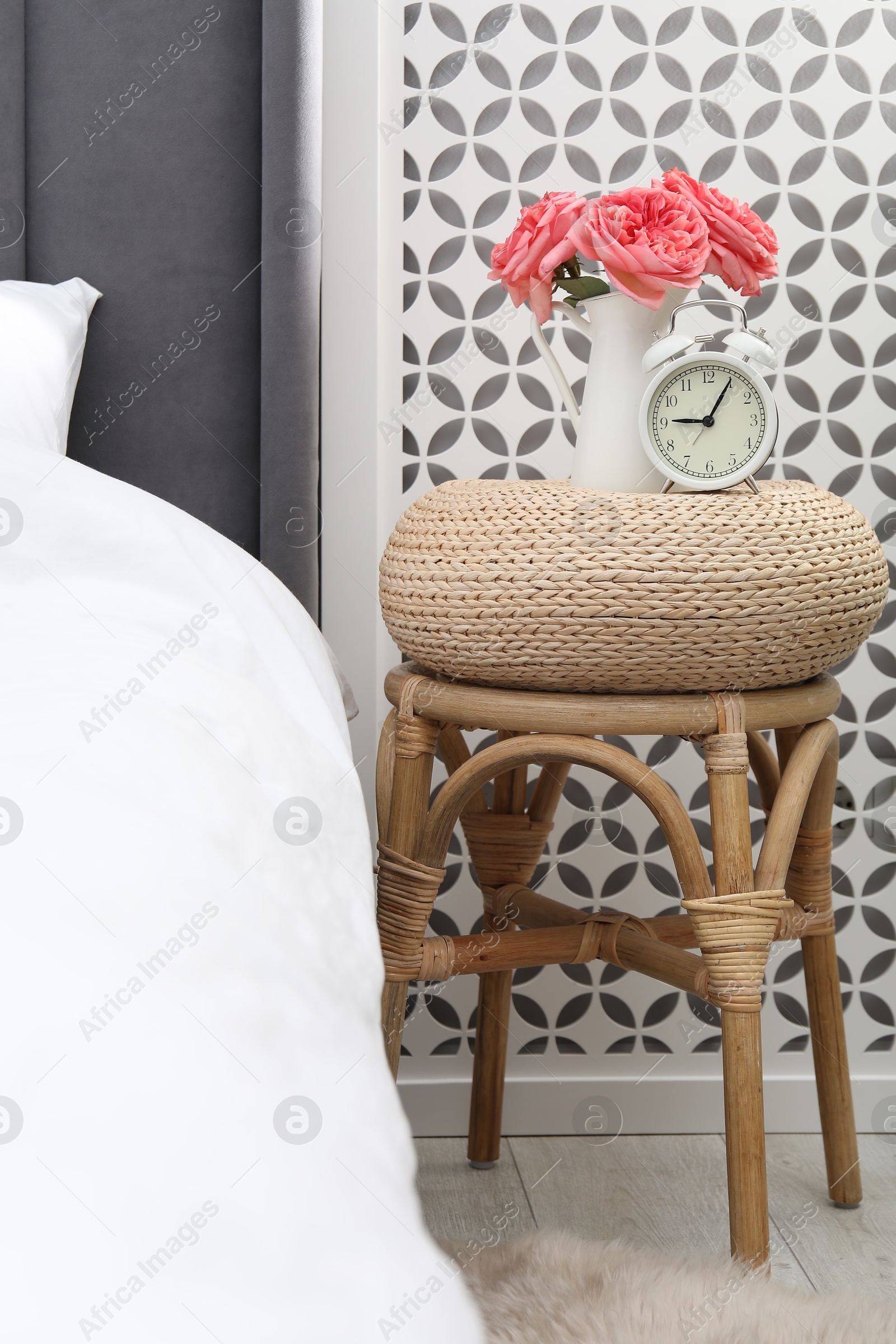 Side table with alarm clock and flowers near bed indoors Photo of Side table with alarm clock and flowers near bed indoors
