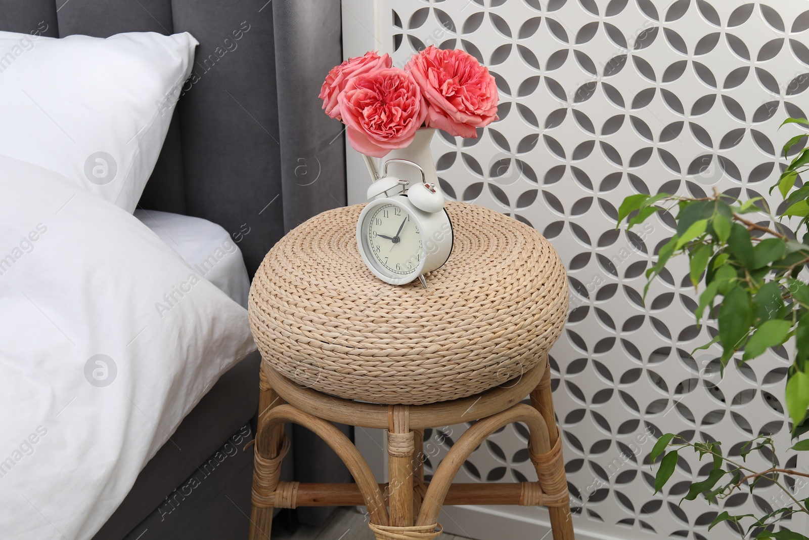 Side table with alarm clock and flowers near bed indoors Photo of Side table with alarm clock and flowers near bed indoors