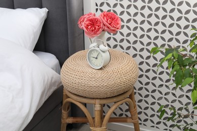 Photo of Side table with alarm clock and flowers near bed indoors