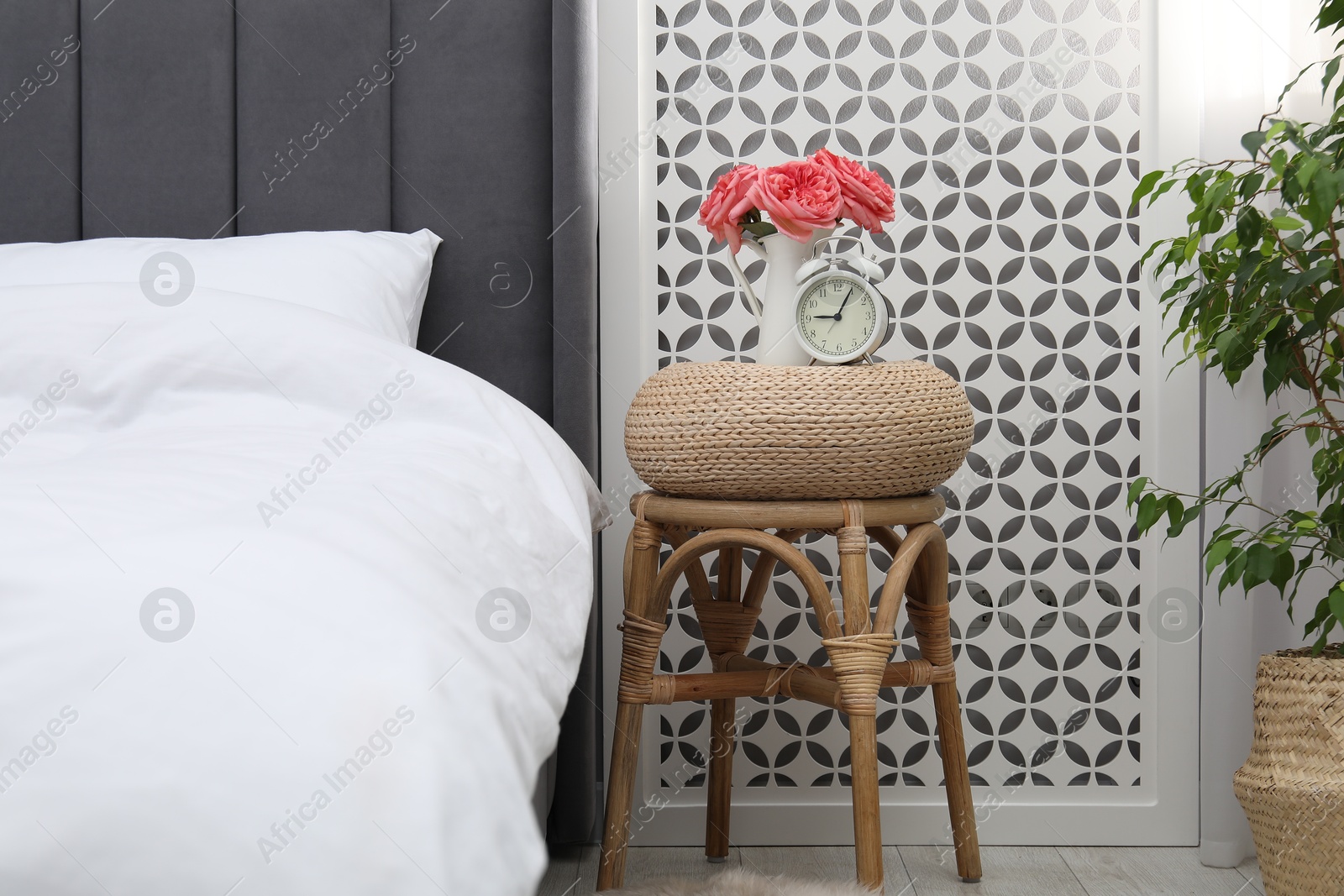 Side table with alarm clock and flowers near bed indoors Photo of Side table with alarm clock and flowers near bed indoors