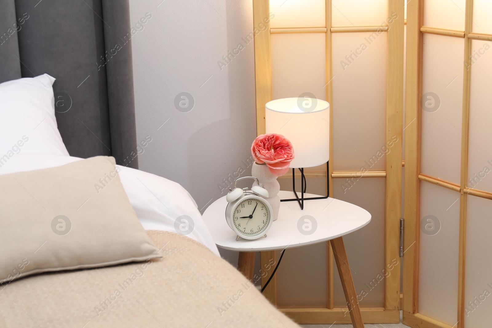 Side table with lamp, alarm clock and flower near bed indoors Photo of Side table with lamp, alarm clock and flower near bed indoors