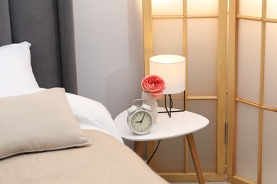 Photo of Side table with lamp, alarm clock and flower near bed indoors