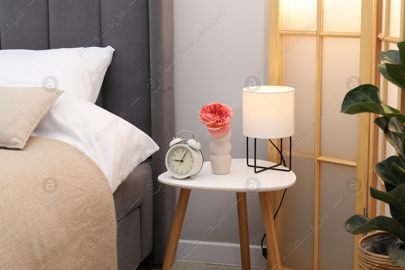 Side table with lamp, alarm clock and flower near bed indoors Photo of Side table with lamp, alarm clock and flower near bed indoors