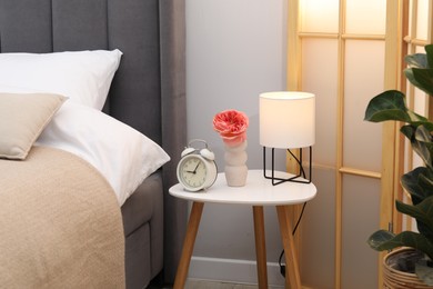 Photo of Side table with lamp, alarm clock and flower near bed indoors