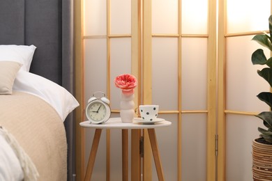 Photo of Side table with alarm clock, cup of tea and flower near bed indoors