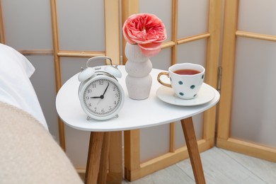 Photo of Side table with alarm clock, cup of tea and flower near bed indoors