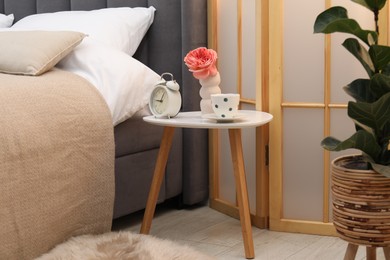 Photo of Side table with alarm clock, cup of tea and flower near bed indoors