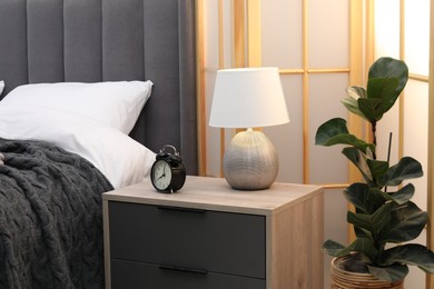 Photo of Bedside table with alarm clock and lamp near bed indoors