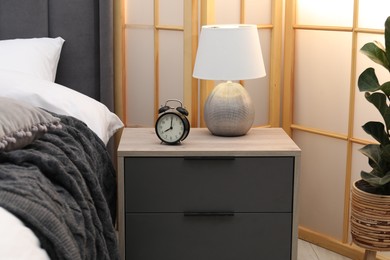 Photo of Bedside table with alarm clock and lamp near bed indoors