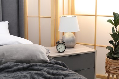 Photo of Bedside table with alarm clock and lamp near bed indoors