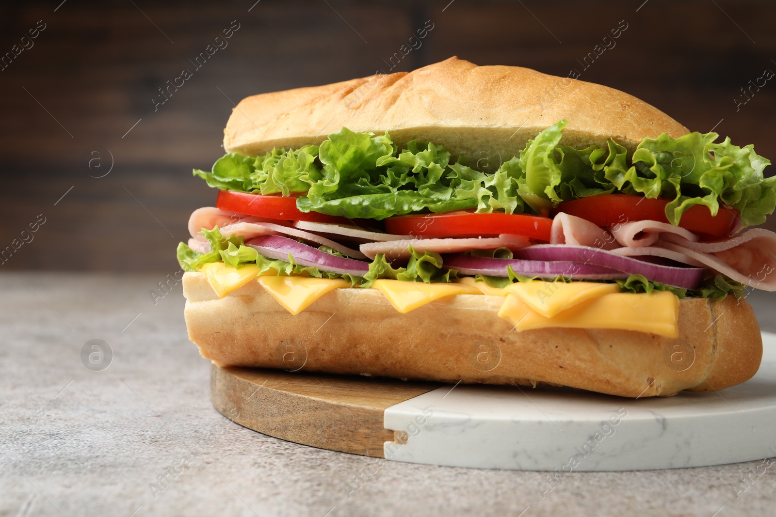 Tasty baguette sandwich with ham, cheese and vegetables on grey textured table against blurred brown background, closeup Photo of Tasty baguette sandwich with ham, cheese and vegetables on grey textured table against blurred brown background, closeup