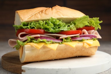 Photo of Tasty baguette sandwich with ham, cheese and vegetables on grey textured table against blurred brown background, closeup