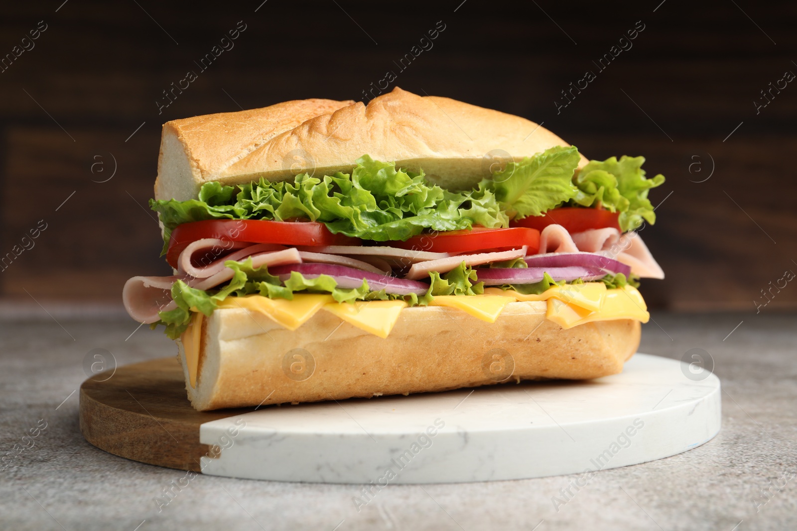 Tasty baguette sandwich with ham, cheese and vegetables on grey textured table against blurred brown background, closeup Photo of Tasty baguette sandwich with ham, cheese and vegetables on grey textured table against blurred brown background, closeup