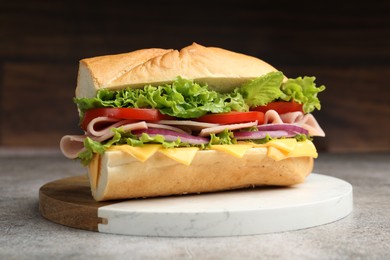 Photo of Tasty baguette sandwich with ham, cheese and vegetables on grey textured table against blurred brown background, closeup