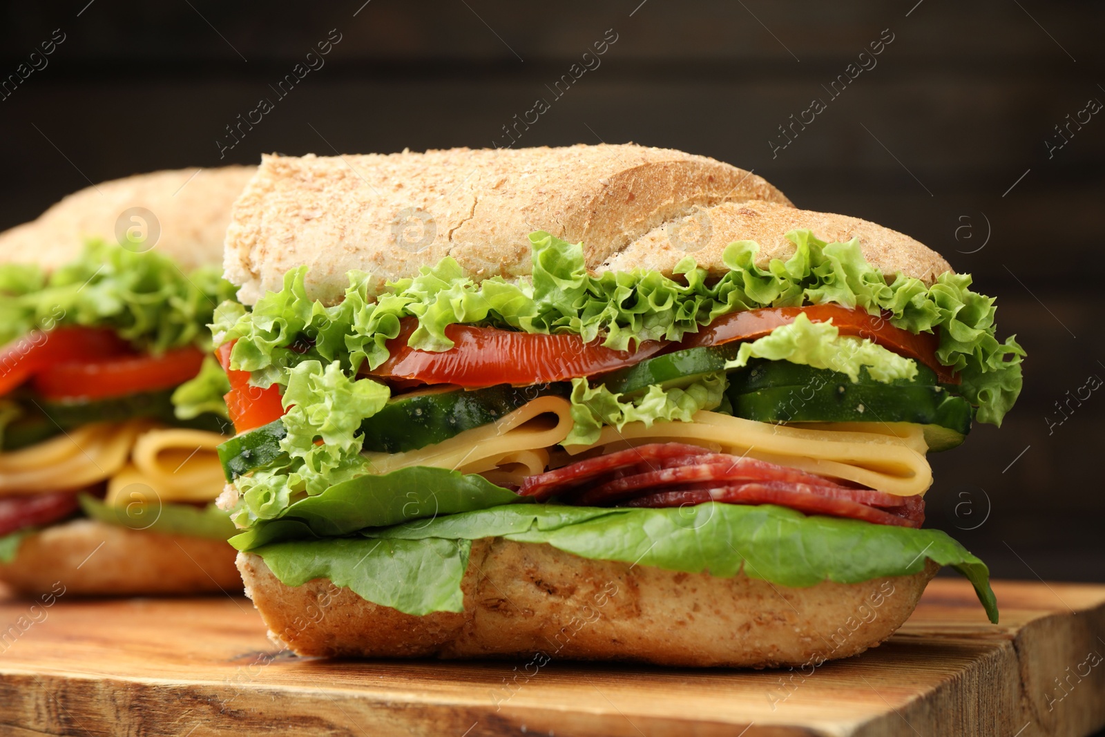 Pieces of tasty baguette sandwich with smoked sausage, cheese and vegetables on wooden board against dark background, closeup Photo of Pieces of tasty baguette sandwich with smoked sausage, cheese and vegetables on wooden board against dark background, closeup