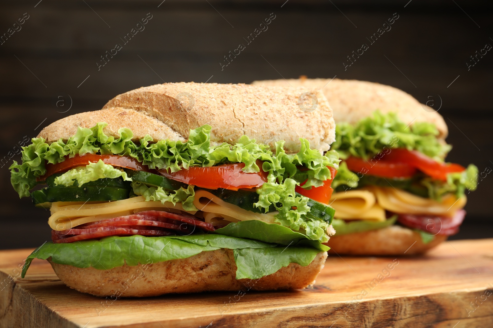 Photo of Pieces of tasty baguette sandwich with smoked sausage, cheese and vegetables on wooden board against dark background, closeup