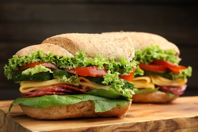Photo of Pieces of tasty baguette sandwich with smoked sausage, cheese and vegetables on wooden board against dark background, closeup
