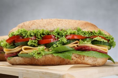 Photo of Tasty baguette sandwich with smoked sausage, cheese and vegetables on wooden board against grey background, closeup