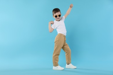 Photo of Happy little boy in sunglasses dancing on light blue background. Space for text