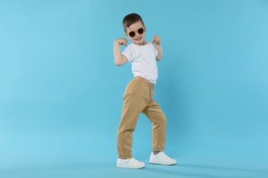 Photo of Happy little boy in sunglasses dancing on light blue background