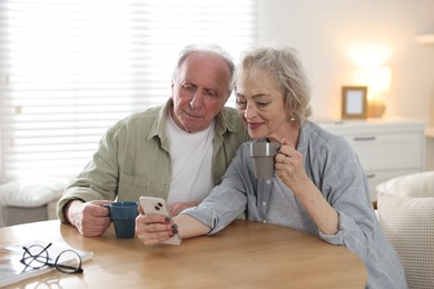 Photo of Lovely senior couple enjoying hot drinks while watching something on smartphone at table indoors