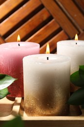 Photo of Burning candles and green leaves on wooden tray, closeup
