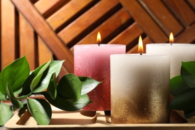 Photo of Burning candles and green leaves on wooden tray, closeup