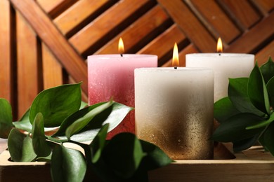 Photo of Burning candles and green leaves on wooden tray, closeup