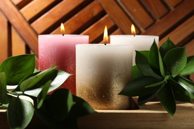 Photo of Burning candles and green leaves on wooden tray, closeup
