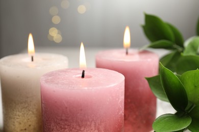 Photo of Burning candles and green leaves on table against blurred lights, closeup