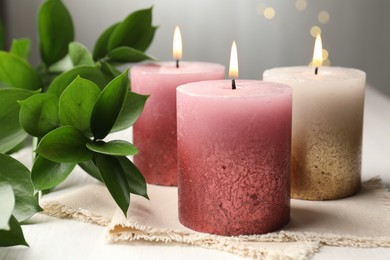Photo of Burning candles and green leaves on white table against blurred lights, closeup