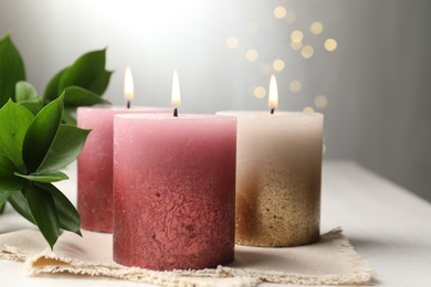Photo of Burning candles and green leaves on white table against blurred lights, closeup