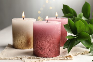 Photo of Burning candles and green leaves on white table against blurred lights, closeup