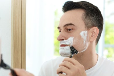 Photo of Handsome man shaving with razor near mirror at home