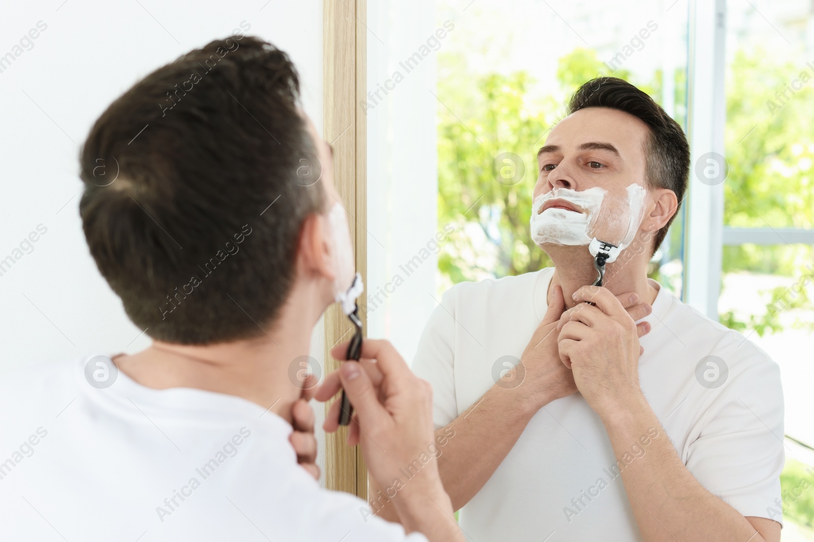 Handsome man shaving with razor near mirror at home Photo of Handsome man shaving with razor near mirror at home