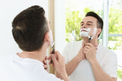Photo of Handsome man shaving with razor near mirror at home