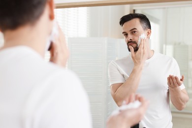 Photo of Handsome man applying shaving foam near mirror in bathroom