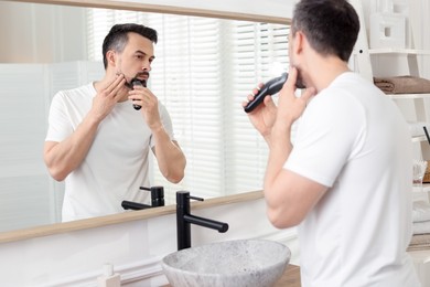 Photo of Handsome man shaving with trimmer near mirror at home